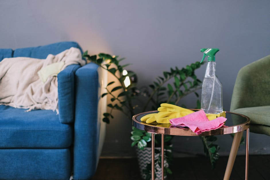 A living room corner featuring a blue fabric sofa with a beige blanket draped over the armrest, adjacent to a round side table with a black top and metallic rose gold legs. On the table, there are yel